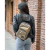 Mochila Hera en color dorado, llevada al hombro por una persona con chaqueta de cuero, en un entorno urbano con paredes de ladrillo.