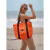 Cartera Melisa de color naranja, con asas negras, llevada por una mujer en la playa.