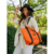 Mujer sonriente con una cartera melisa de color naranja, usando un sombrero negro y blusa de rayas, en un entorno natural.