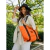 Mujer sonriente con una cartera melisa de color naranja, usando un sombrero negro y blusa de rayas, en un entorno natural.