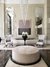 Área de estar com decoração elegante, incluindo duas poltronas brancas, uma mesa redonda preta com um arranjo de orquídeas brancas e um grande lustre pendente de crist comprar lustres, luminarias e pendentes na Lustres Gênesisal.