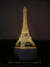 Abajur de LED em formato de Torre Eiffel aceso, ideal para leitura e decoração de quartos ou escritórios. Lustres Gênesis Site online venda de Lustres e Iluminação