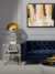 Abajur Delfos Dourado com Cristal em sala de estar moderna, ao lado de sofá azul e mesa lateral com livros.   Lustres Gênesis Site online venda de Lustres e Iluminação