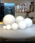 Globos de vidro fosco leitoso de diferentes tamanhos exibidos em uma mesa, com decoração de fundo, da linha Jabuticaba. Lustres Gênesis loja online de Lustres e Iluminação Decorativa