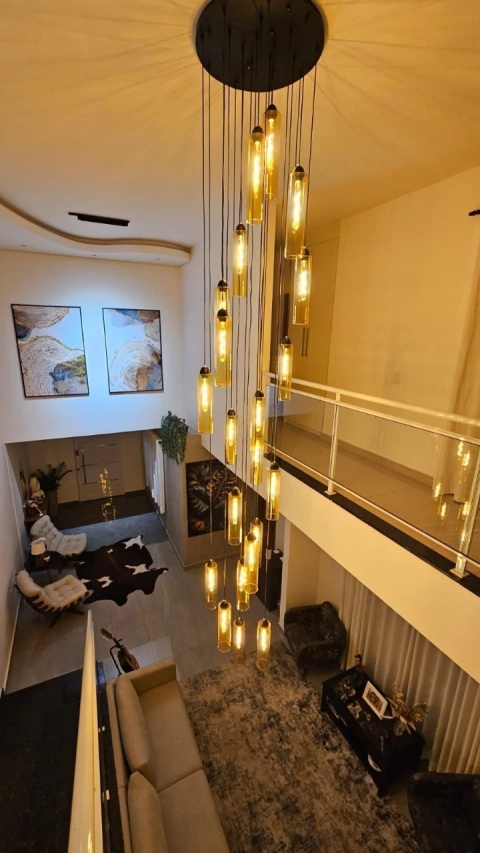 Lustre Pendente Moderno Dunáh com 21 tubos âmbar em base redonda, instalado em casa com pé direito duplo, iluminando sala de estar sofisticada.