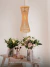 Luminária pendente de rattan com detalhes em madeira, suspensa sobre uma mesa decorada com flores. Lustres Gênesis Site online venda de Lustres e Iluminação