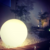 Luminária de chão esfera Soleil branca em um jardim com pedras brancas e plantas ao fundo. Lustres Gênesis loja online de Lustres e Iluminação Decorativa