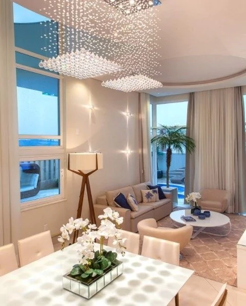 Interior de uma sala de estar moderna com decoração elegante, incluindo um lustre de cristal, sofá bege com almofadas azuis, mesa de centro branca e plantas decorativas. Lustres Gênesis loja online de Lustres e Iluminação Decorativa