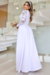 Vestido longo branco em piquet com manga longa e detalhes em renda, modelo Denise, exibido por uma mulher segurando um buquê de flores secas em um ambiente elegante.