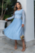 Modelo usando um vestido plissado em crepe azul claro com renda nas mangas, segurando uma bolsa dourada em um ambiente externo