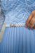 Modelo posando com vestido longo plissado em crepe azul claro, com detalhes em renda nas mangas, em ambiente elegante com paredes de mármore.