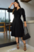 Modelo usando um vestido midi plissado preto em suede com mangas longas, posando em um ambiente moderno.