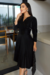 Modelo usando um vestido midi plissado preto em suede com mangas longas, posando em um ambiente moderno.