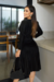 Modelo usando um vestido midi plissado preto em suede com mangas longas, posando em um ambiente moderno.