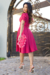 Modelo usando um vestido midi pink com mangas bufantes e detalhes em malha lese, segurando uma bolsa de flores, em um ambiente externo.