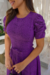 Modelo usando um vestido midi de malha lese na cor roxa, com mangas bufantes e cinto na cintura, em um ambiente iluminado.
