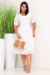 Mulher usando um vestido midi branco rendado com botões e mangas curtas, segurando uma bolsa de palha, ao lado de plantas verdes em vasos brancos.