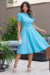 Mulher usando um vestido midi em malha lese azul, com mangas bufantes, em um ambiente externo com plantas ao fundo.