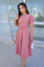Modelo vestindo um vestido midi rosa de malha lese com mangas bufantes e cinto, segurando uma bolsa pequena