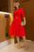 Modelo usando um vestido midi vermelho de malha lese forrado, com mangas 3/4 e cinto. Ela segura uma bolsa dourada e está em um ambiente interno com paredes de madeira.
