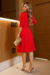 Modelo usando um vestido midi vermelho de malha lese forrado, com mangas 3/4 e cinto. Ela segura uma bolsa dourada e está em um ambiente interno com paredes de madeira.