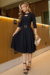 Vestido midi de malha com manga 3/4, cor preta, detalhe em recorte na gola, cinto na cintura e saia rodada, usado por uma modelo em ambiente elegante.