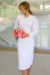 Modelo usando um vestido tubinho branco de malha Lese com detalhes em elastano, zíper nas costas e mangas bufantes, segurando um buquê de flores em um ambiente moderno.