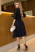 Vestido midi de malha com manga 3/4, cor preta, detalhe em recorte na gola, cinto na cintura e saia rodada, usado por uma modelo em ambiente elegante.