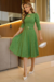Modelo usando um vestido midi verde de manga 3/4, com detalhes em malha lese e cinto, em um ambiente elegante.