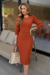 Modelo vestindo um vestido tubinho de malha lese com elastano na cor terracota, com zíper nas costas, posando em um ambiente moderno.