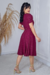 Mulher usando vestido midi malha lese na cor vinho, com detalhes em babados e um cinto na cintura, posando em ambiente claro.