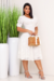 Mulher usando um vestido midi branco rendado com botões e mangas curtas, segurando uma bolsa de palha, ao lado de plantas verdes em vasos brancos.