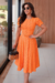 Modelo usando vestido midi laranja de malha Lese, com detalhes recortados e mangas bufantes, segurando uma bolsa.