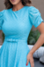 Mulher usando um vestido midi em malha lese azul, com mangas bufantes, em um ambiente externo com plantas ao fundo.