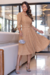 Vestido midi em tom nude com mangas 3/4 e detalhe na cintura, usado por uma mulher em ambiente elegante.