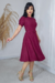 Mulher usando vestido midi malha lese na cor vinho, com detalhes em babados e um cinto na cintura, posando em ambiente claro.