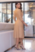 Vestido midi em tom nude com mangas 3/4 e detalhe na cintura, usado por uma mulher em ambiente elegante.