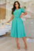 Vestido midi em malha lese na cor verde água, com mangas curtas e detalhe de laço na cintura, combinado com sapatos nude e uma bolsa colorida.