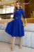 Vestido midi azul com manga 3/4 e detalhe de fivela na cintura, modelo elegante em ambiente contemporâneo.