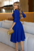 Vestido midi azul com manga 3/4 e detalhe de fivela na cintura, modelo elegante em ambiente contemporâneo.