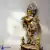Estátua de Krishna em pose de flauta, adornada em detalhes dourados, com figuras menores ao lado, sobre base ornamental.