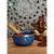 Tigela tibetana azul com detalhes em dourado, acompanhada de um bastão, sobre uma mesa de madeira. Ao fundo, há uma figura de Ganesha e uma decoração em estilo mandala.