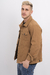 Hombre posando con una chaqueta gabardina de color camel, de perfil, con fondo blanco.