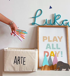 Contenedor rectangular de tela color crudo con la palabra ARTE impresa, junto a una mano sosteniendo marcadores de colores pastel y decoración infantil en estante