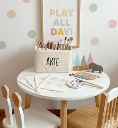 Contenedor rectangular de tela con la palabra "ARTE" impresa, lleno de pinceles y lápices de colores sobre una mesa blanca redonda en un cuarto infantil decorado con papeles de dibujo y decoración de pared de colores pastel.