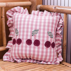 Almofada decorativa Vichy Cerejinhas com estampa xadrez rosa claro, babados nas bordas e bordados de cerejinhas vermelhas, acompanhada por outros itens de decoração infantil e um ursinho de tecido.