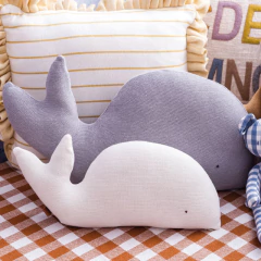 Almofada de tricô em formato de baleia, na cor cinza, decorando ambiente infantil sobre toalha xadrez marrom e branco, com outras almofadas ao fundo.