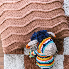 Almofada de tricô quadrada 45x45cm com textura de ondas em tom bege, acompanhada de um bichinho de pelúcia colorido, ideal para decoração de ambientes infantis.