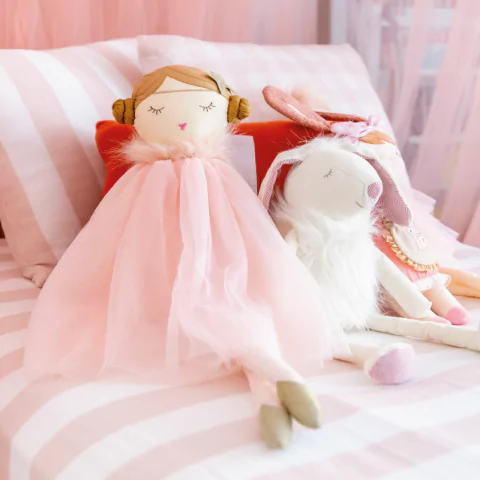 Almofada retangular de tricô listrada 30 x 60cm em tom rosa, decorando cama infantil com bonecas de pano e coelho de pelúcia, criando ambiente aconchegante para quarto de bebê.