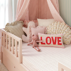 Almofada retangular Vichy com babado e sianinha sobre cama infantil decorada, acompanhada de outras almofadas coloridas e ursinho de tecido.